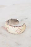 A Louis Vuitton cuff bracelet in beige monogram canvas with silver-tone metal studs and a silver interior, resting on a light marble surface.