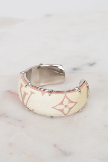 A Louis Vuitton cuff bracelet in beige monogram canvas with silver-tone metal studs and a silver interior, resting on a light marble surface.