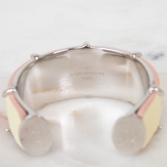 A Louis Vuitton cuff bracelet with a silver-tone metal band, featuring pale yellow and light pink enamel inlays, resting on a white marble surface.