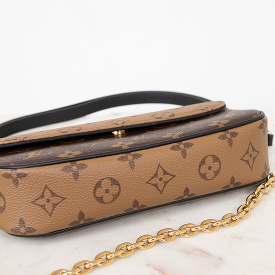 A Louis Vuitton monogram canvas shoulder bag in tan and brown, featuring a black leather strap and a gold-tone chain link detail, displayed on a white marble surface against a white background.