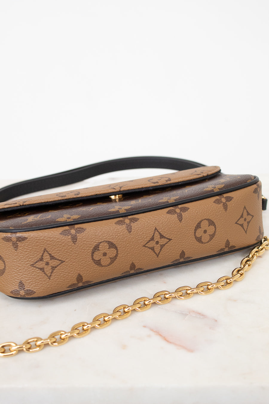 A Louis Vuitton monogram canvas shoulder bag in tan and brown, featuring a black leather strap and a gold-tone chain link detail, displayed on a white marble surface against a white background.