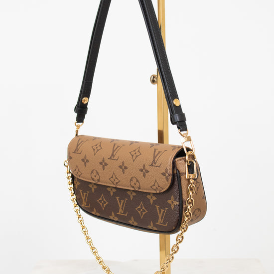 Louis Vuitton monogram shoulder bag in brown coated canvas with a black leather shoulder strap and gold-tone chain detail, displayed on a gold stand against a white background.