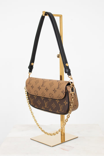 Louis Vuitton monogram shoulder bag in brown coated canvas with a black leather shoulder strap and gold-tone chain detail, displayed on a gold stand against a white background.