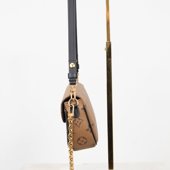 A Louis Vuitton monogram shoulder bag in brown coated canvas with a black leather strap and gold-tone chain, displayed on a gold metal stand against a white background.