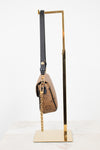 A Louis Vuitton monogram shoulder bag in brown coated canvas with a black leather strap and gold-tone chain, displayed on a gold metal stand against a white background.