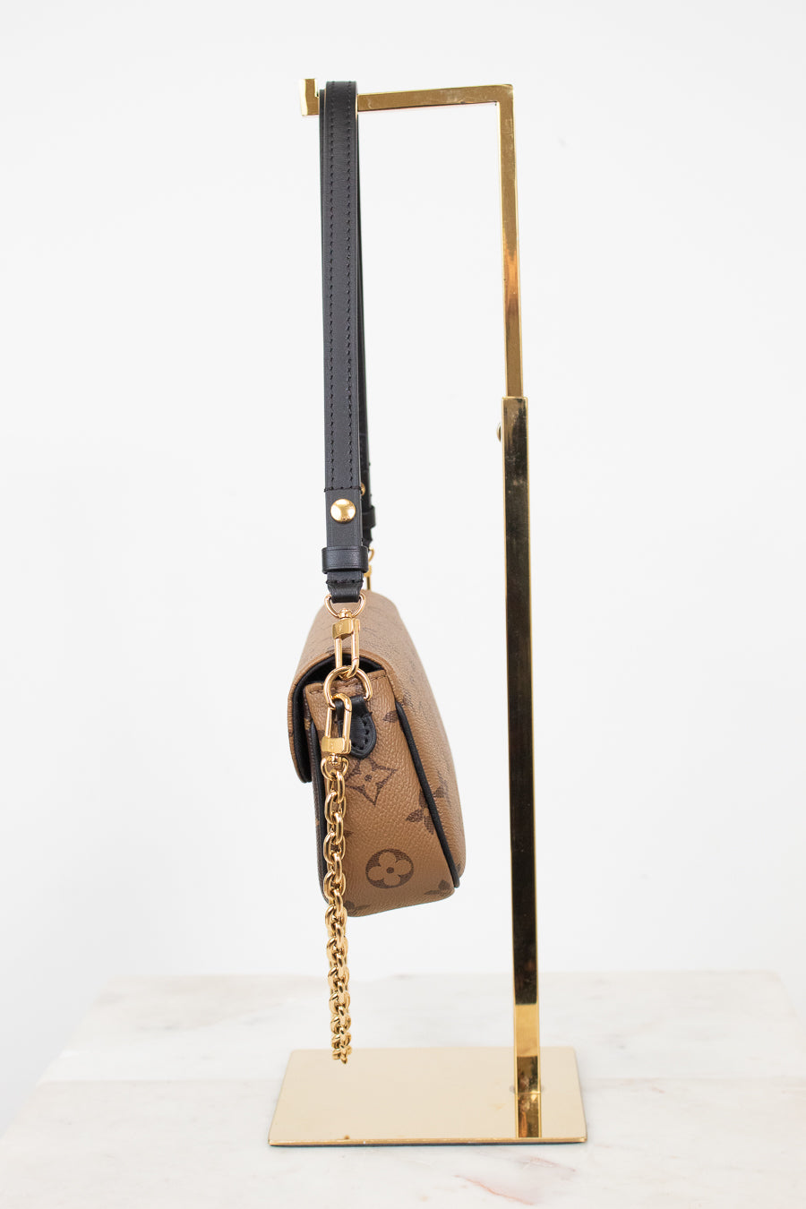 A Louis Vuitton monogram shoulder bag in brown coated canvas with a black leather strap and gold-tone chain, displayed on a gold metal stand against a white background.