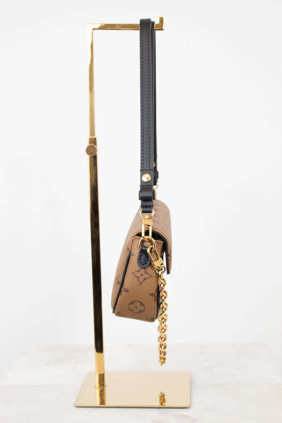A Louis Vuitton monogram shoulder bag in brown coated canvas with a black leather strap and gold-tone chain, displayed on a gold metal stand against a white background.