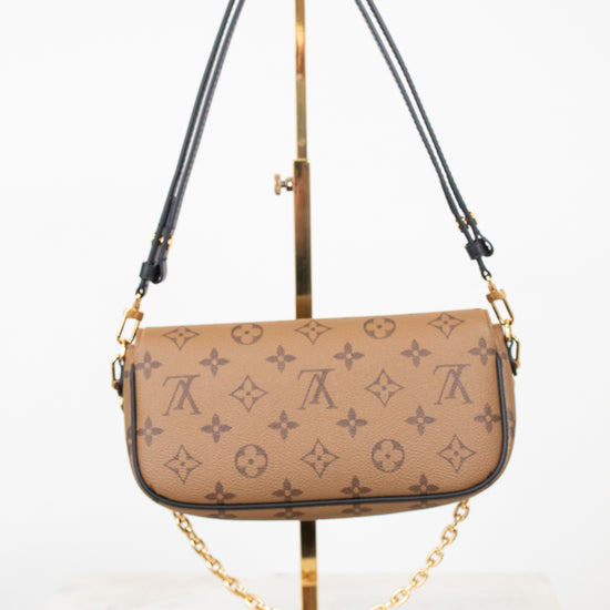 A Louis Vuitton monogram shoulder bag in beige coated canvas with dark brown leather trim and a gold-tone chain strap, displayed on a gold stand against a white background.