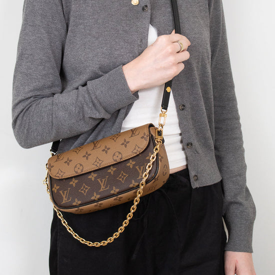A woman wearing a gray cardigan, white t-shirt, and black pants carries a brown Louis Vuitton monogram shoulder bag with a gold chain strap.