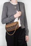 A woman wearing a gray cardigan, white t-shirt, and black pants carries a brown Louis Vuitton monogram shoulder bag with a gold chain strap.