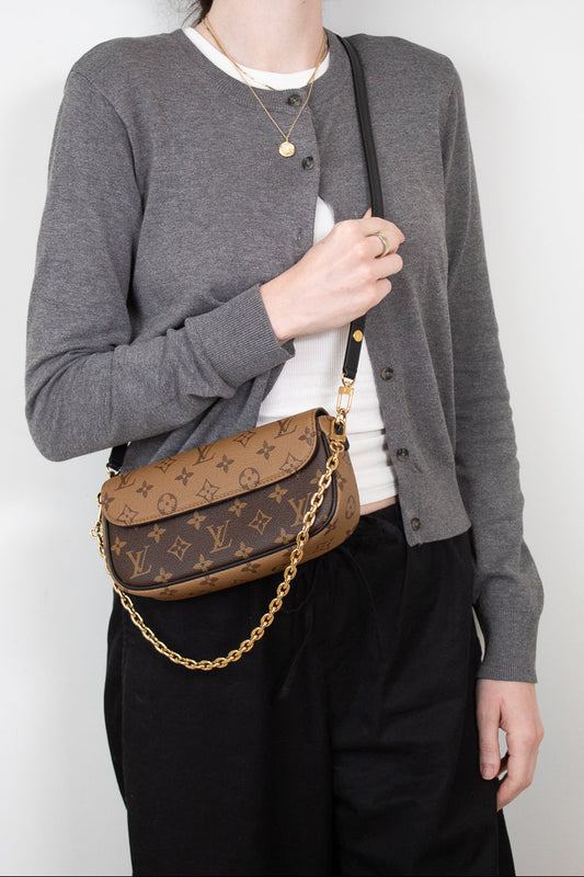 A woman wearing a gray cardigan, white t-shirt, and black pants carries a brown Louis Vuitton monogram shoulder bag with a gold chain strap.