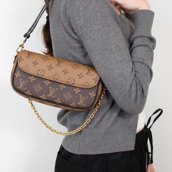 A woman wearing a gray long-sleeve top and black pants carries a Louis Vuitton monogram shoulder bag with a gold chain strap against a plain white background.