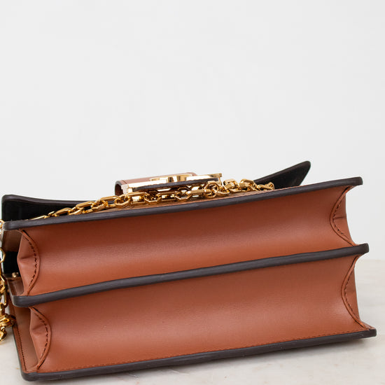 A close-up of a brown leather book-style handbag with black trim and a gold chain strap, resting on a light marble surface against a white background.