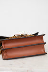 A close-up of a brown leather book-style handbag with black trim and a gold chain strap, resting on a light marble surface against a white background.