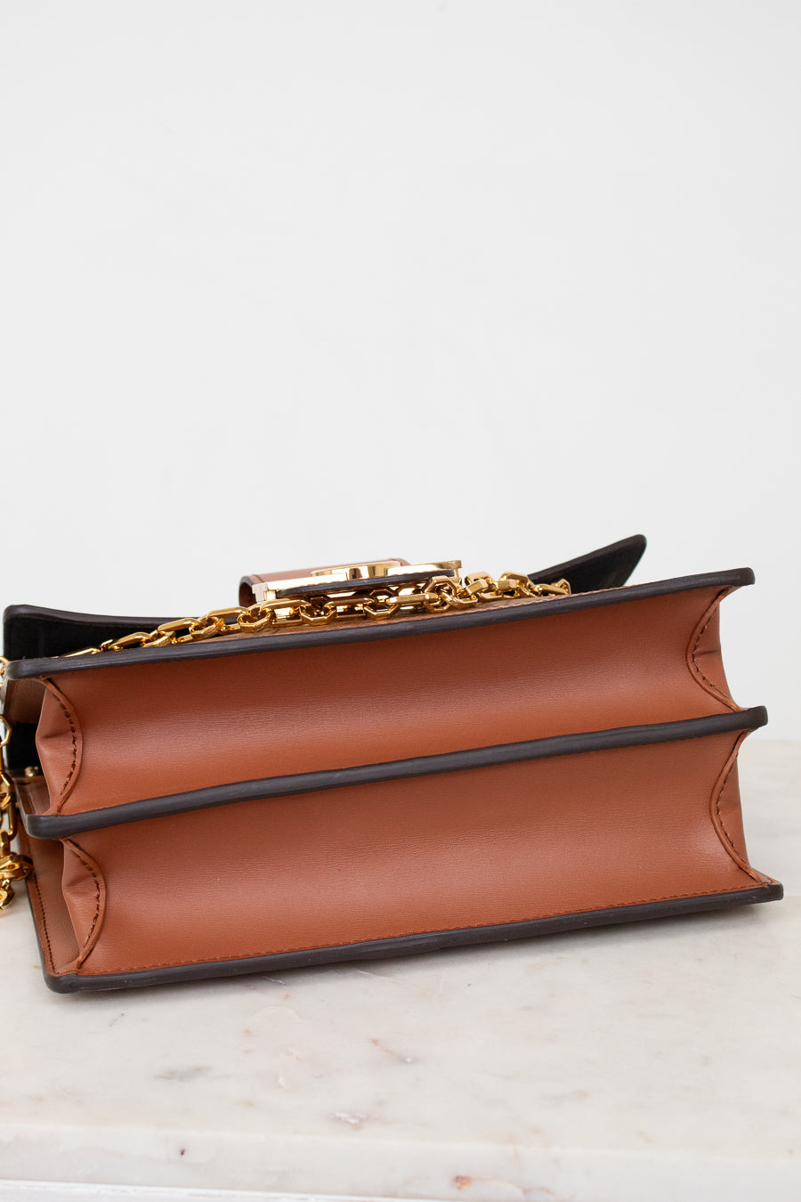 A close-up of a brown leather book-style handbag with black trim and a gold chain strap, resting on a light marble surface against a white background.