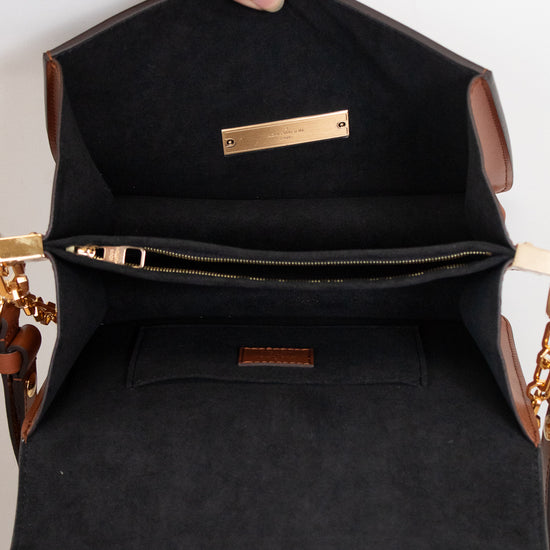 An open black handbag with brown leather trim, showing a black interior lining, a zippered pocket, and gold-tone hardware. A hand with rings is holding the bag open against a plain white background.