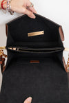 An open black handbag with brown leather trim, showing a black interior lining, a zippered pocket, and gold-tone hardware. A hand with rings is holding the bag open against a plain white background.