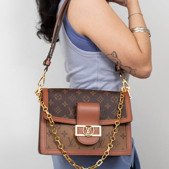 A woman wearing a light blue top and skirt carries a Louis Vuitton monogram shoulder bag with a brown leather flap, gold-tone hardware, and a gold chain strap against a plain light gray background.