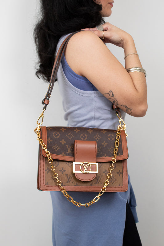 A woman wearing a light blue top and skirt carries a Louis Vuitton monogram shoulder bag with a brown leather flap, gold-tone hardware, and a gold chain strap against a plain light gray background.