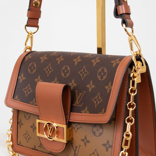 A Louis Vuitton shoulder bag in brown monogram coated canvas with tan leather trim and a gold-tone chain strap, displayed on a stand against a light background.