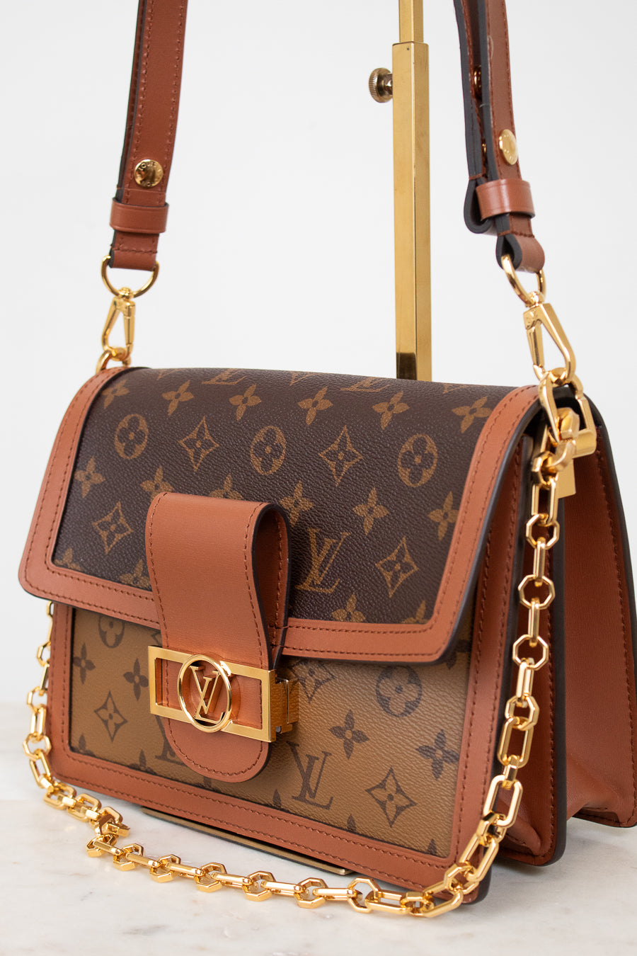 A Louis Vuitton shoulder bag in brown monogram coated canvas with tan leather trim and a gold-tone chain strap, displayed on a stand against a light background.