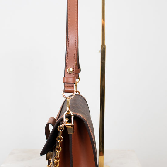 A brown leather shoulder bag with gold-tone hardware and a gold chain strap, displayed on a white marble surface with a gold metal stand against a white background.