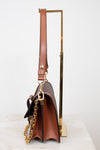 A brown leather shoulder bag with gold-tone hardware and a gold chain strap, displayed on a white marble surface with a gold metal stand against a white background.
