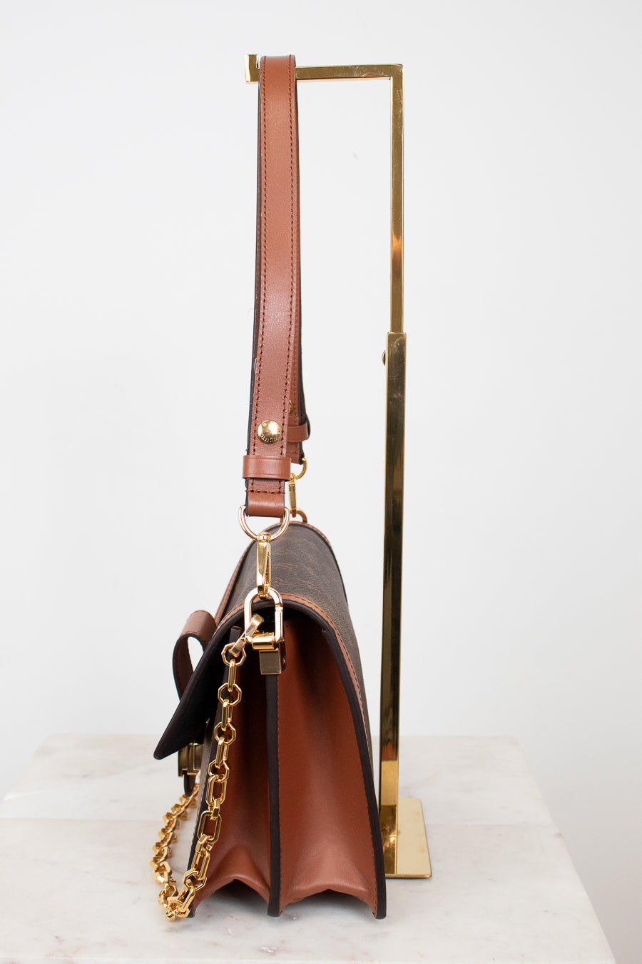 A brown leather shoulder bag with gold-tone hardware and a gold chain strap, displayed on a white marble surface with a gold metal stand against a white background.