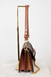 A brown leather shoulder bag with gold-tone hardware, displayed on a gold metal stand against a white background.