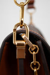 Close-up of a Louis Vuitton handbag hardware detail, showing a gold-tone metal clasp and chain with the Louis Vuitton name engraved on a small tag, attached to a dark brown leather bag against a light gray background.