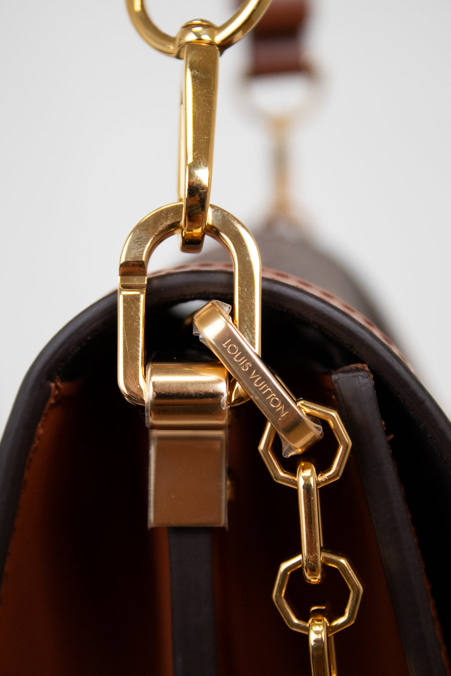Close-up of a Louis Vuitton handbag hardware detail, showing a gold-tone metal clasp and chain with the Louis Vuitton name engraved on a small tag, attached to a dark brown leather bag against a light gray background.
