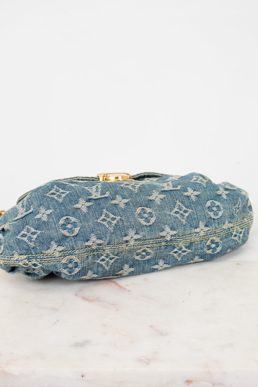 A Louis Vuitton monogrammed denim bag viewed from the bottom with a gold-tone clasp, resting on a light marble surface against a white background.