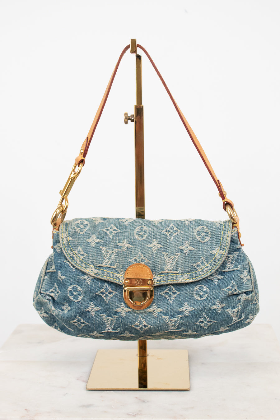 A Louis Vuitton monogram denim shoulder bag in blue, displayed on a gold metal stand against a white background.