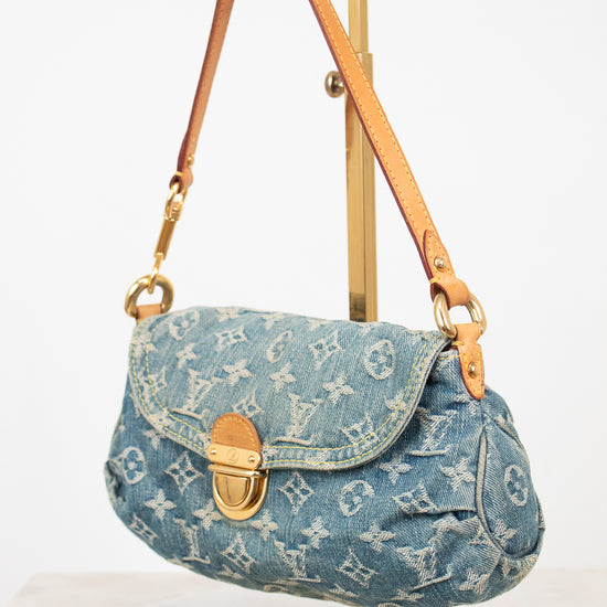 A Louis Vuitton monogram canvas shoulder bag in light blue denim, displayed on a gold metal stand against a white background.