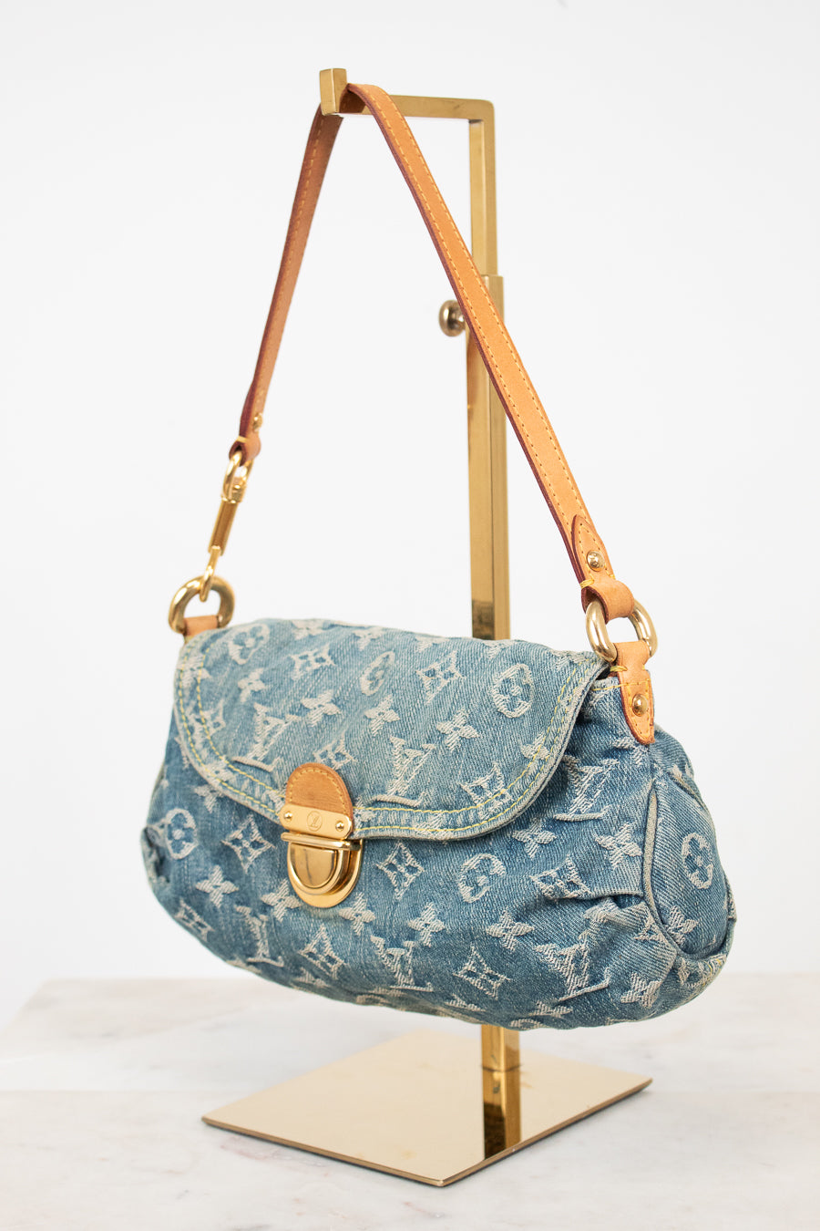 A Louis Vuitton monogram canvas shoulder bag in light blue denim, displayed on a gold metal stand against a white background.