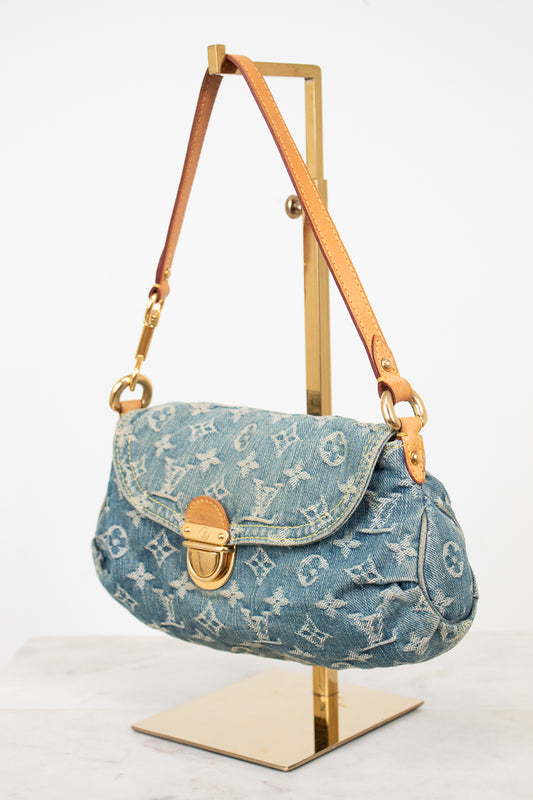 A Louis Vuitton monogram canvas shoulder bag in light blue denim, displayed on a gold metal stand against a white background.
