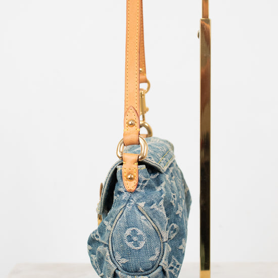 A Louis Vuitton monogrammed blue denim shoulder bag with tan leather trim and gold-tone hardware, hanging from a gold metal display stand against a white background.