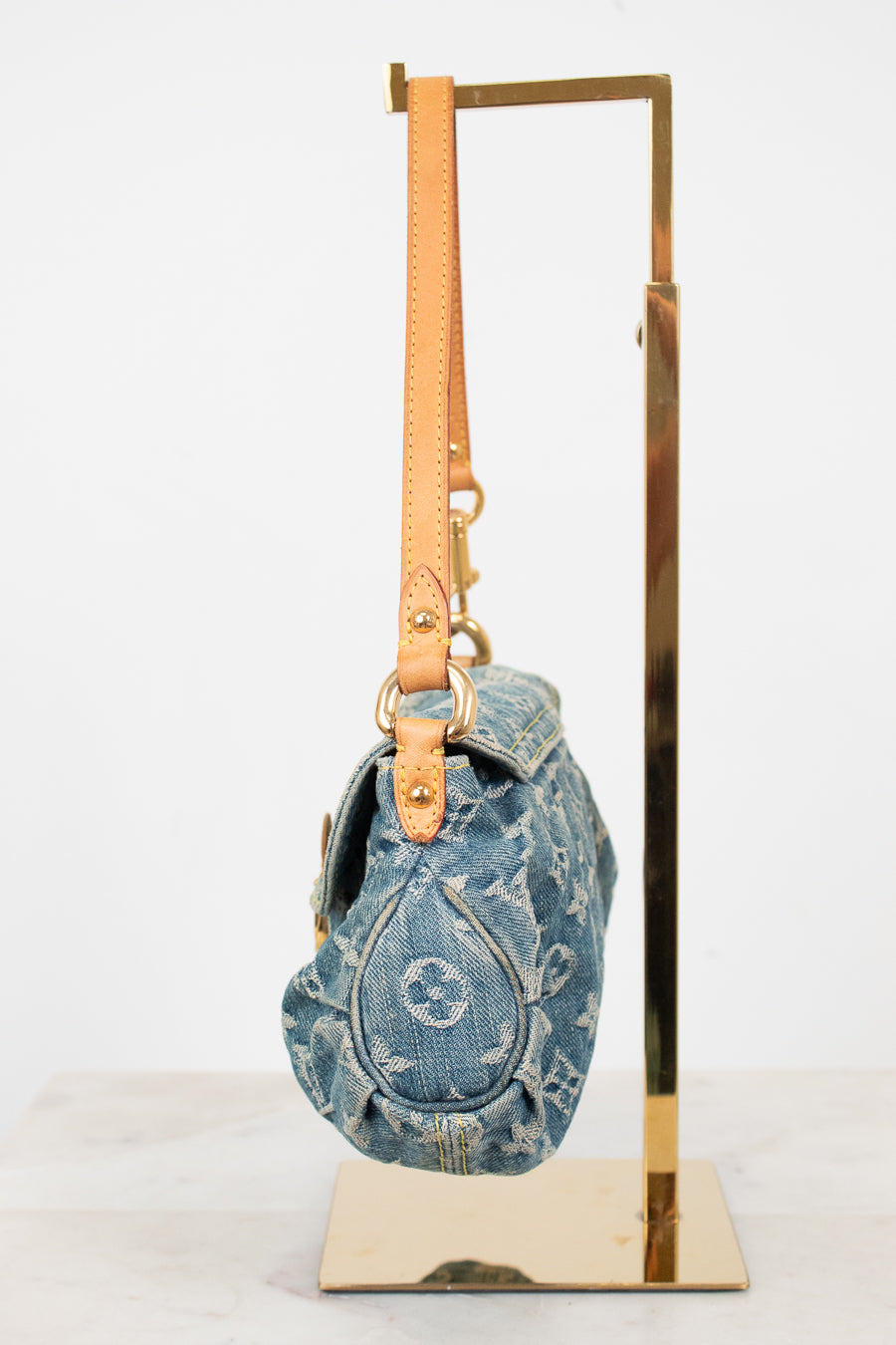 A Louis Vuitton monogrammed blue denim shoulder bag with tan leather trim and gold-tone hardware, hanging from a gold metal display stand against a white background.