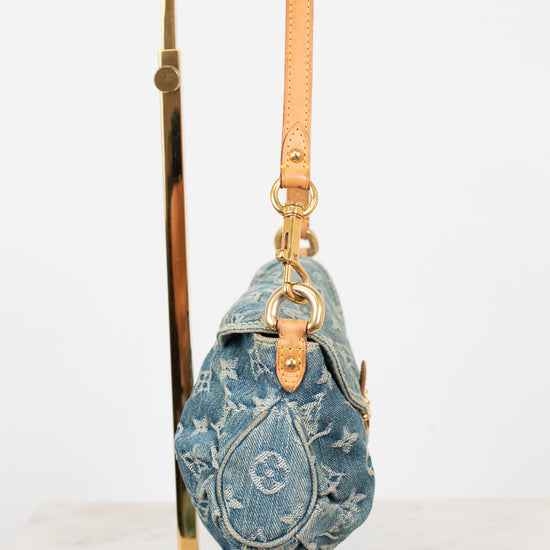 A blue denim shoulder bag with a tan leather strap and gold-tone hardware, displayed on a gold metal stand against a white background.