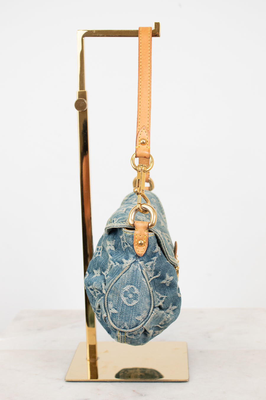A blue denim shoulder bag with a tan leather strap and gold-tone hardware, displayed on a gold metal stand against a white background.