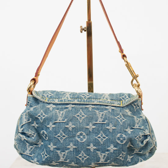 A Louis Vuitton denim shoulder bag with the brand's signature monogram pattern, displayed on a gold-colored stand against a white background.