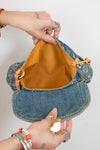 A denim shoulder bag with a mustard yellow interior lining, held open by two hands against a plain white background.