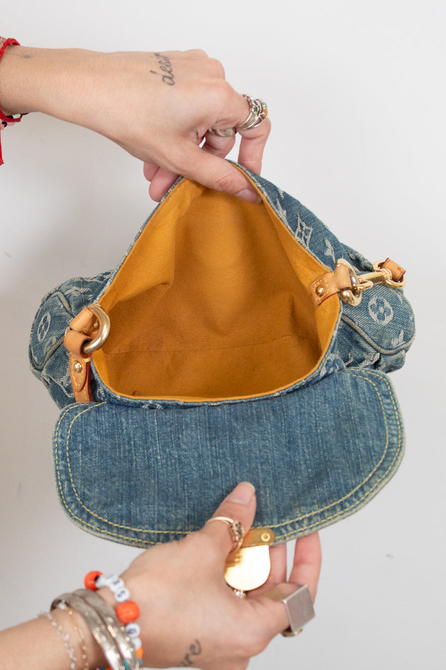 A denim shoulder bag with a mustard yellow interior lining, held open by two hands against a plain white background.