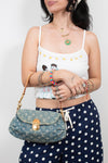 A woman wearing a white graphic camisole and navy polka-dot pants carries a blue denim monogram shoulder bag with gold hardware.