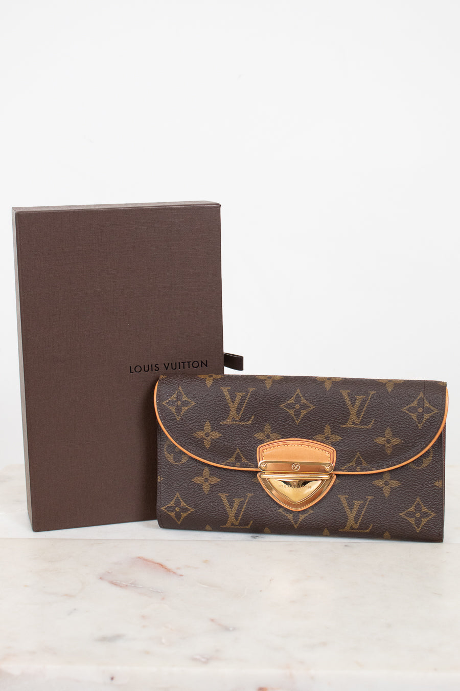 Louis Vuitton monogram wallet in brown coated canvas with gold-tone hardware, shown next to its brown presentation box on a light marble surface against a white background.