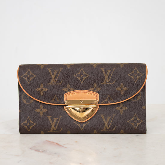 A Louis Vuitton monogram wallet in dark brown coated canvas with tan leather trim and a gold-tone push-lock closure, displayed on a light marble surface against a white background.