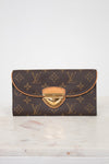 A Louis Vuitton monogram wallet in dark brown coated canvas with tan leather trim and a gold-tone push-lock closure, displayed on a light marble surface against a white background.
