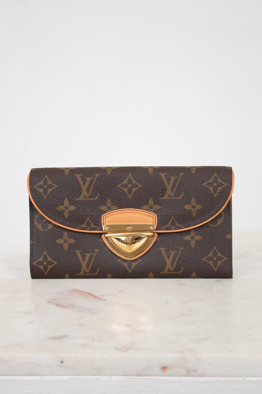 A Louis Vuitton monogram wallet in dark brown coated canvas with tan leather trim and a gold-tone push-lock closure, displayed on a light marble surface against a white background.