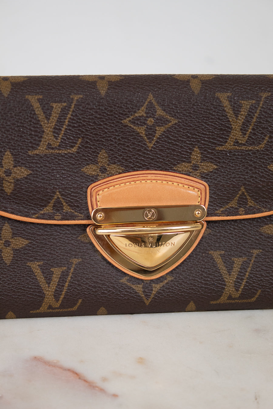 Louis Vuitton monogram canvas clutch with a tan leather strap and a gold-tone metal clasp on a light marble surface.