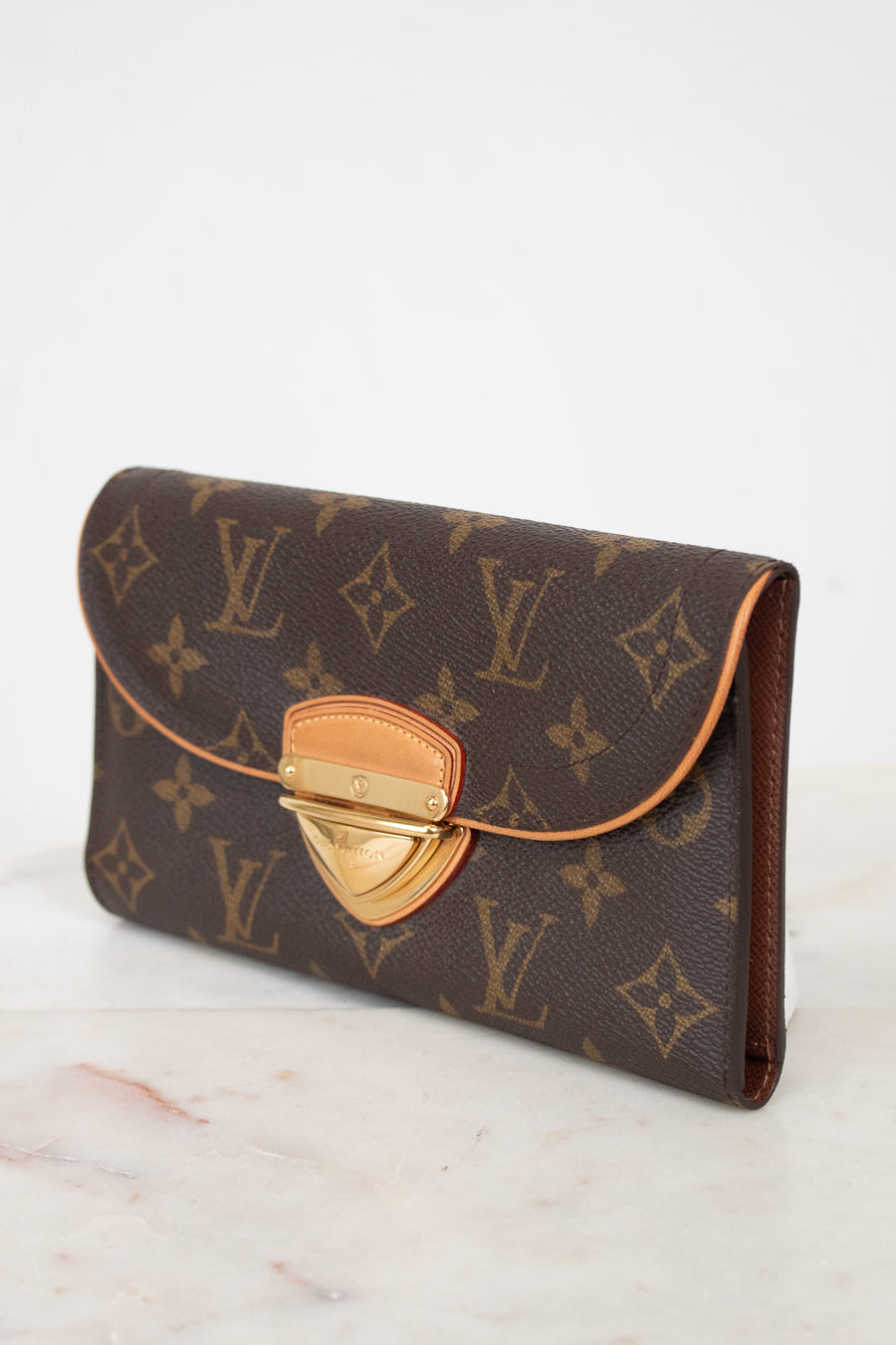 A Louis Vuitton monogram coated canvas clutch wallet with a brown coated canvas flap and gold-tone metal clasp, resting on a light marble surface against a white background.